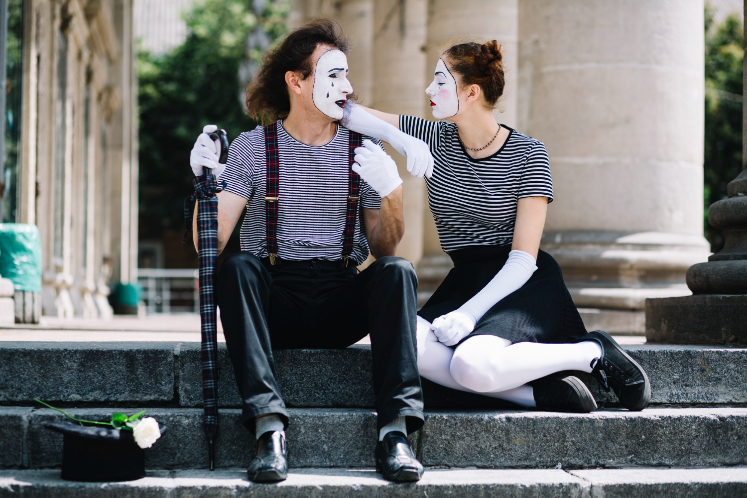 Casal de artistas encena em espaço público, sentados no degrau de um monumento em uma praça.